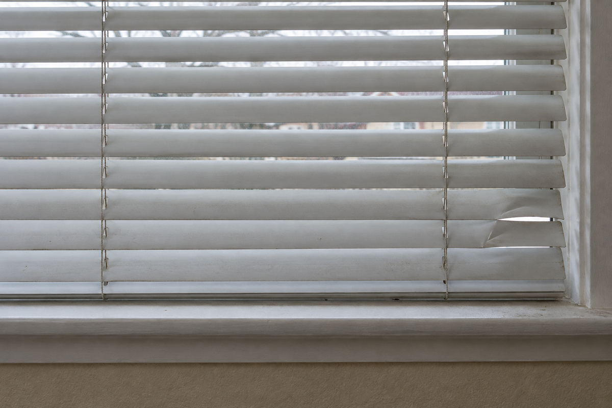 Worn white window blind with two bent lower slats leaving an uneven gap